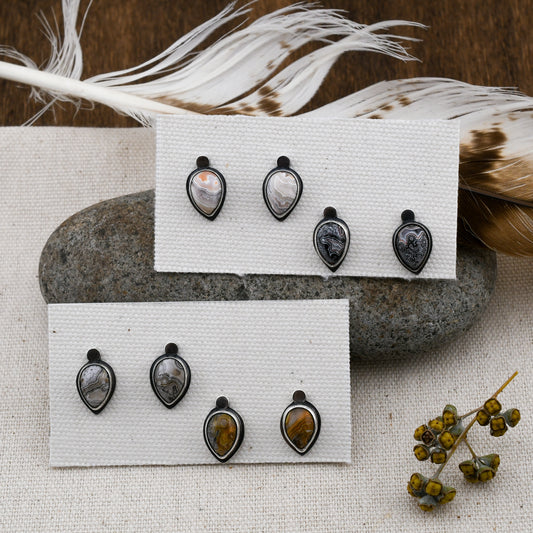Four pairs of sterling silver teardrop-shaped stud earrings with crazy lace agate, displayed on white textured cards over a gray stone with feathers and dried flowers in the background.