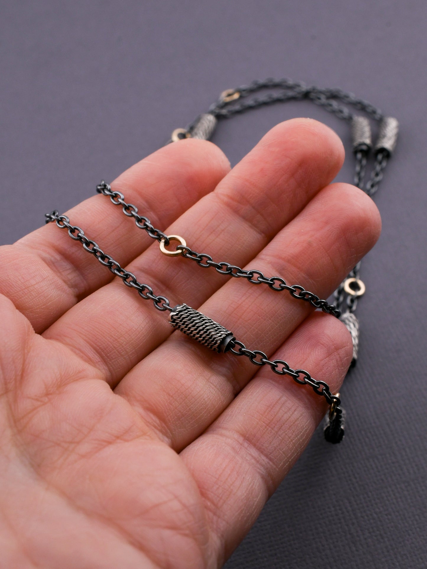 Close-up of a hand holding a black chain necklace and woven silver beads with 14k gold links against a dark background