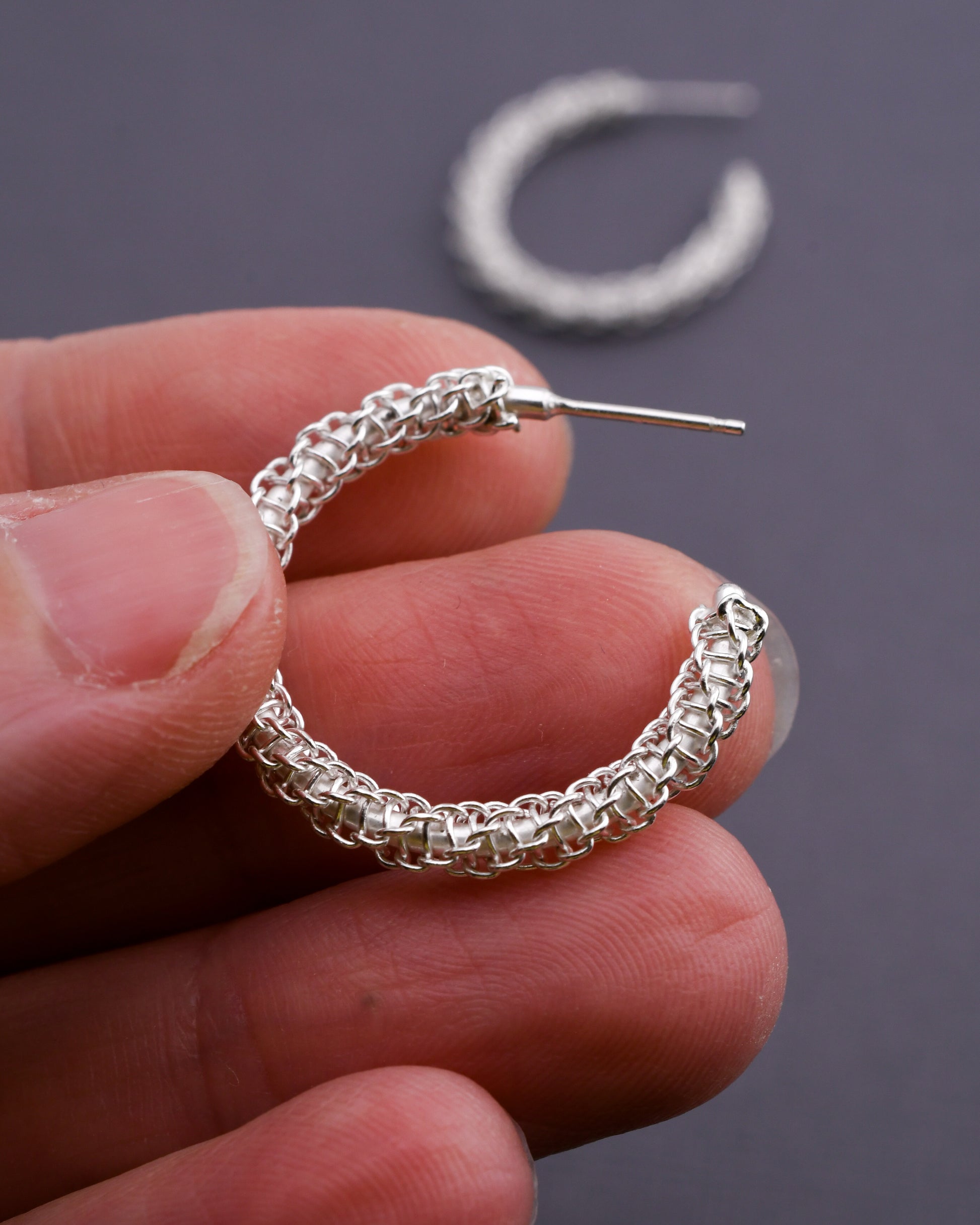 Woven silver hoop earrings held in a hand with a dark background