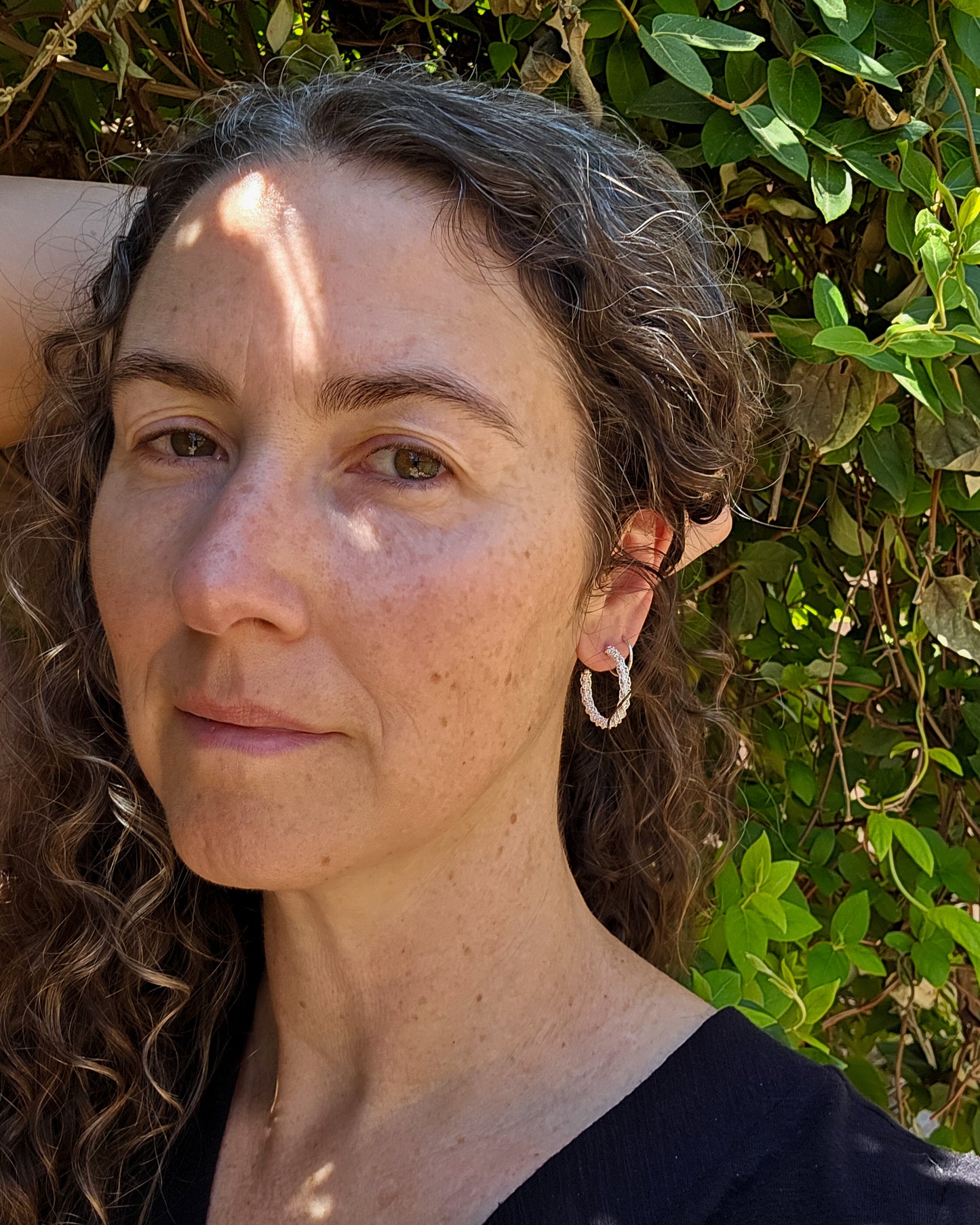 Woven silver Sinnet hoop earrings shown worn on a person in front of a leafy background