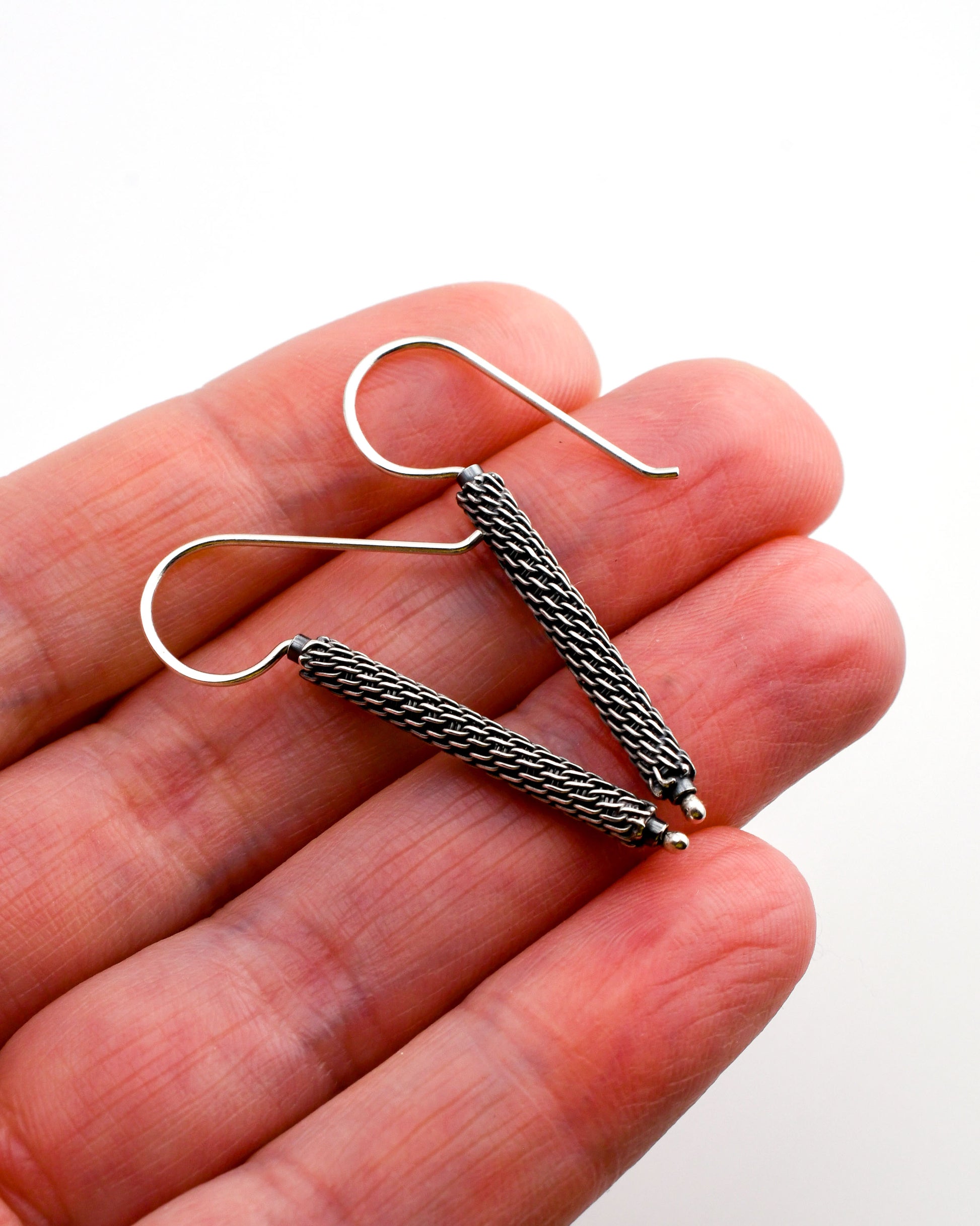 Woven oxidized silver column earrings held in a hand over a white background