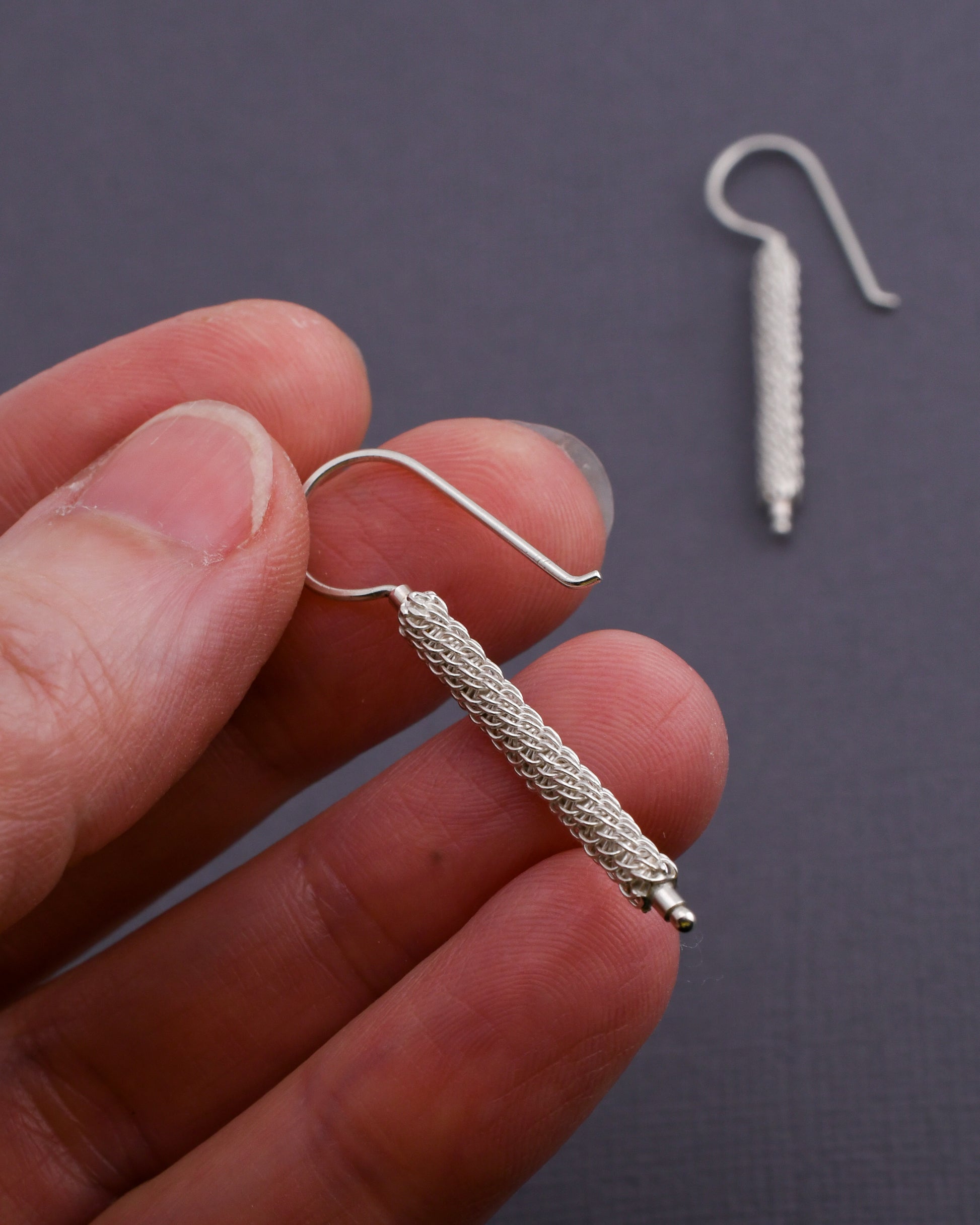 Woven silver column earrings held in a hand over a dark background