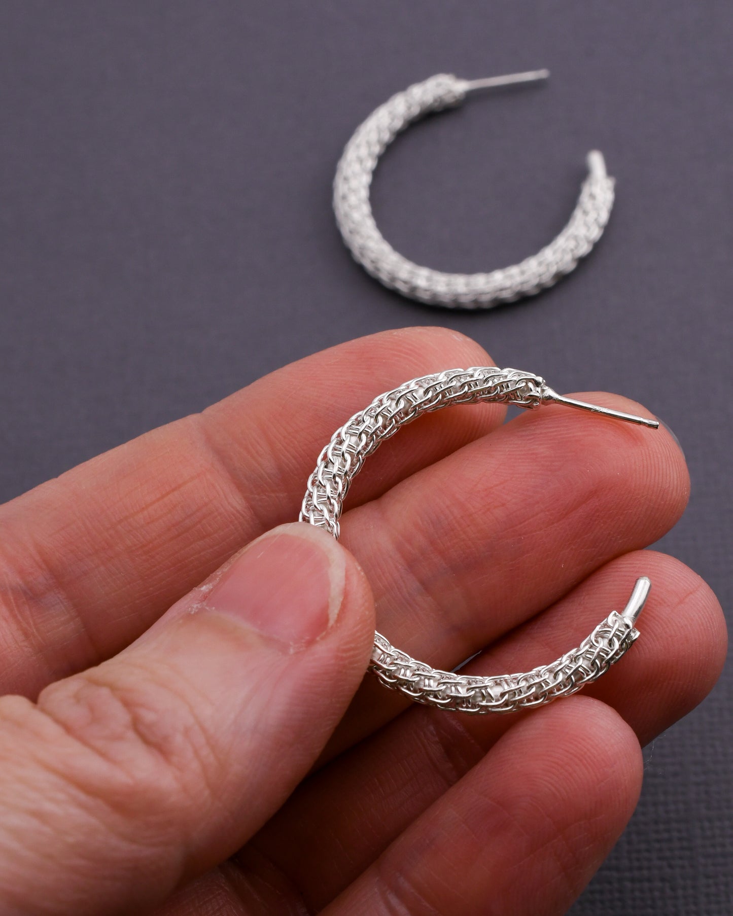 Woven silver hoop earrings held in a hand with a dark background