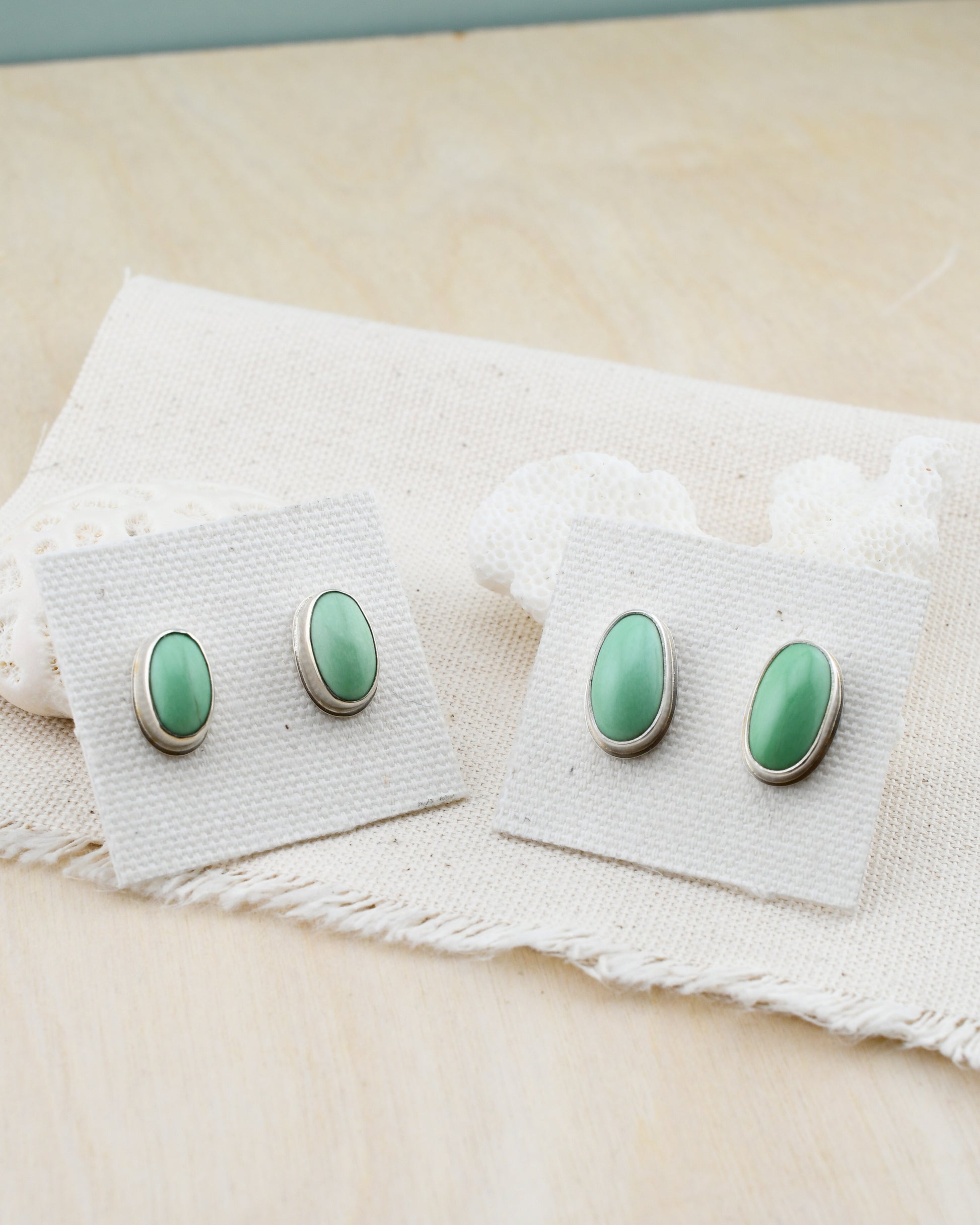 two pairs of silver stud earrings with oval green variscite on linen cards propped against white coral and a light wood background
