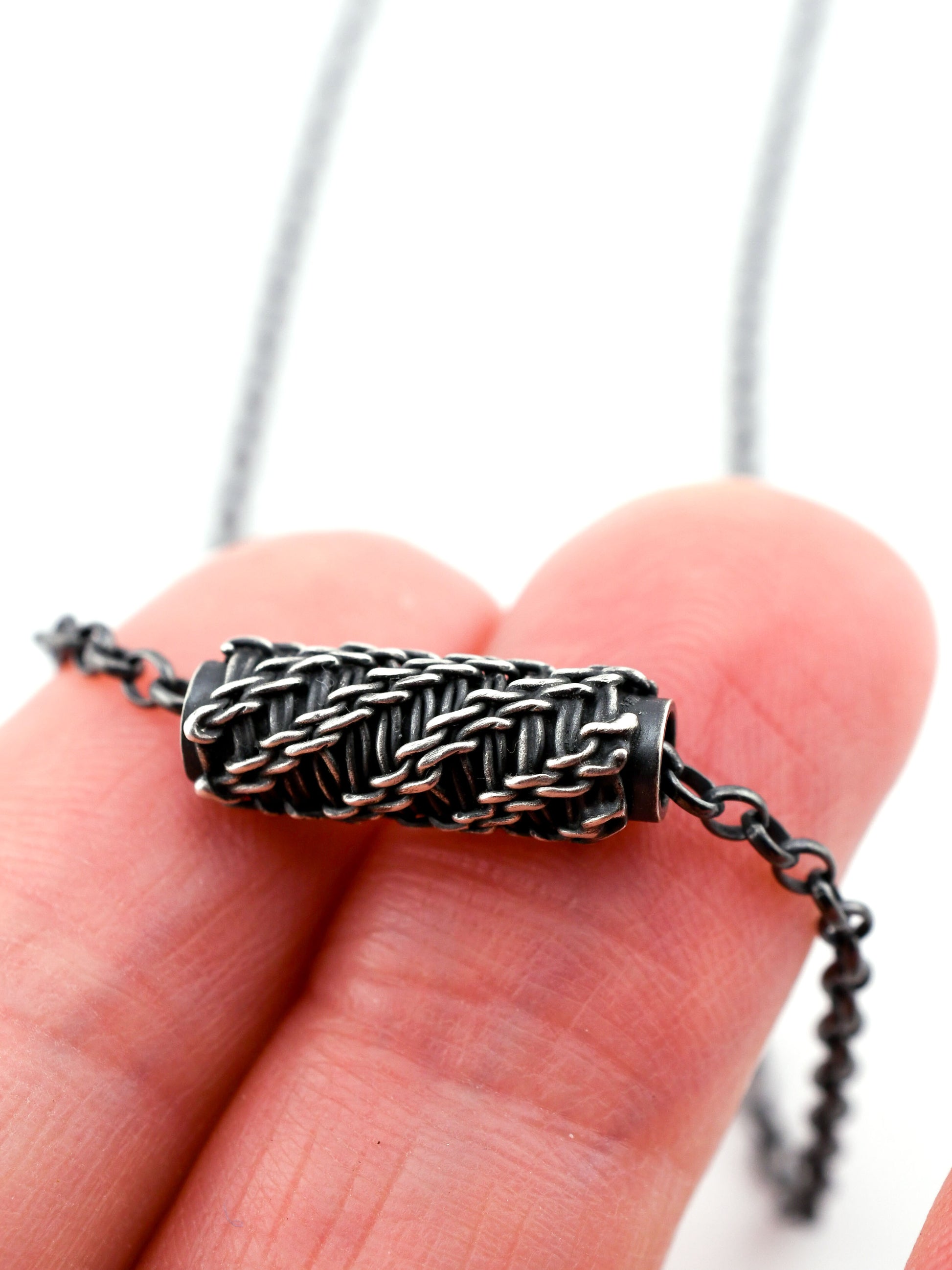 Close-up of a hand holding a silver necklace with a woven pendant against a white background