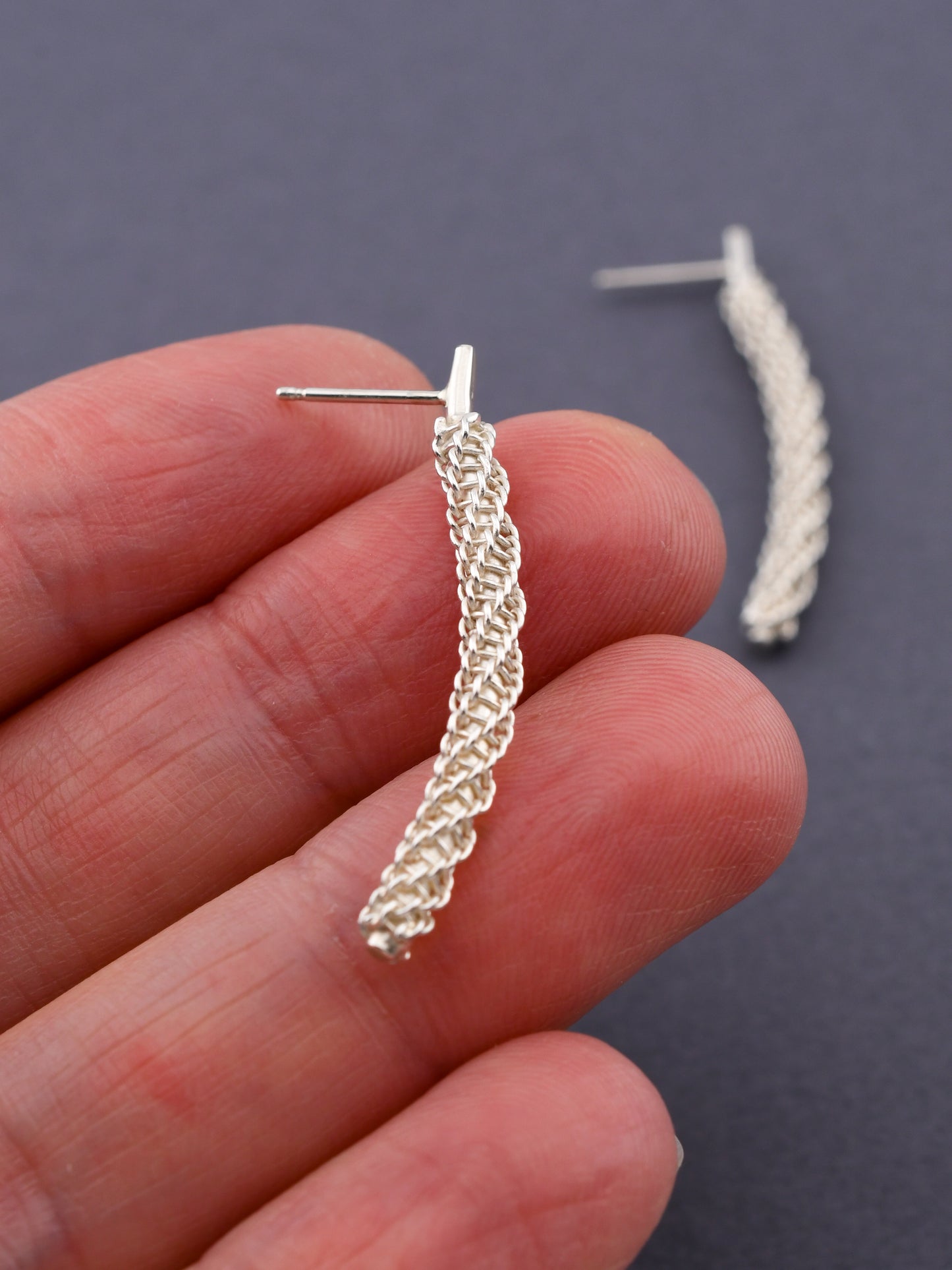 Silver braided earring held between fingers against a dark background
