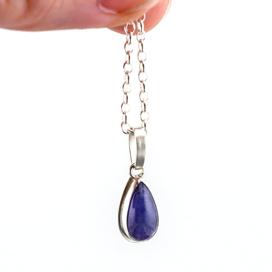 A teardrop shaped Tanzanite pendant hangs from a silver chain held between fingers over a white background