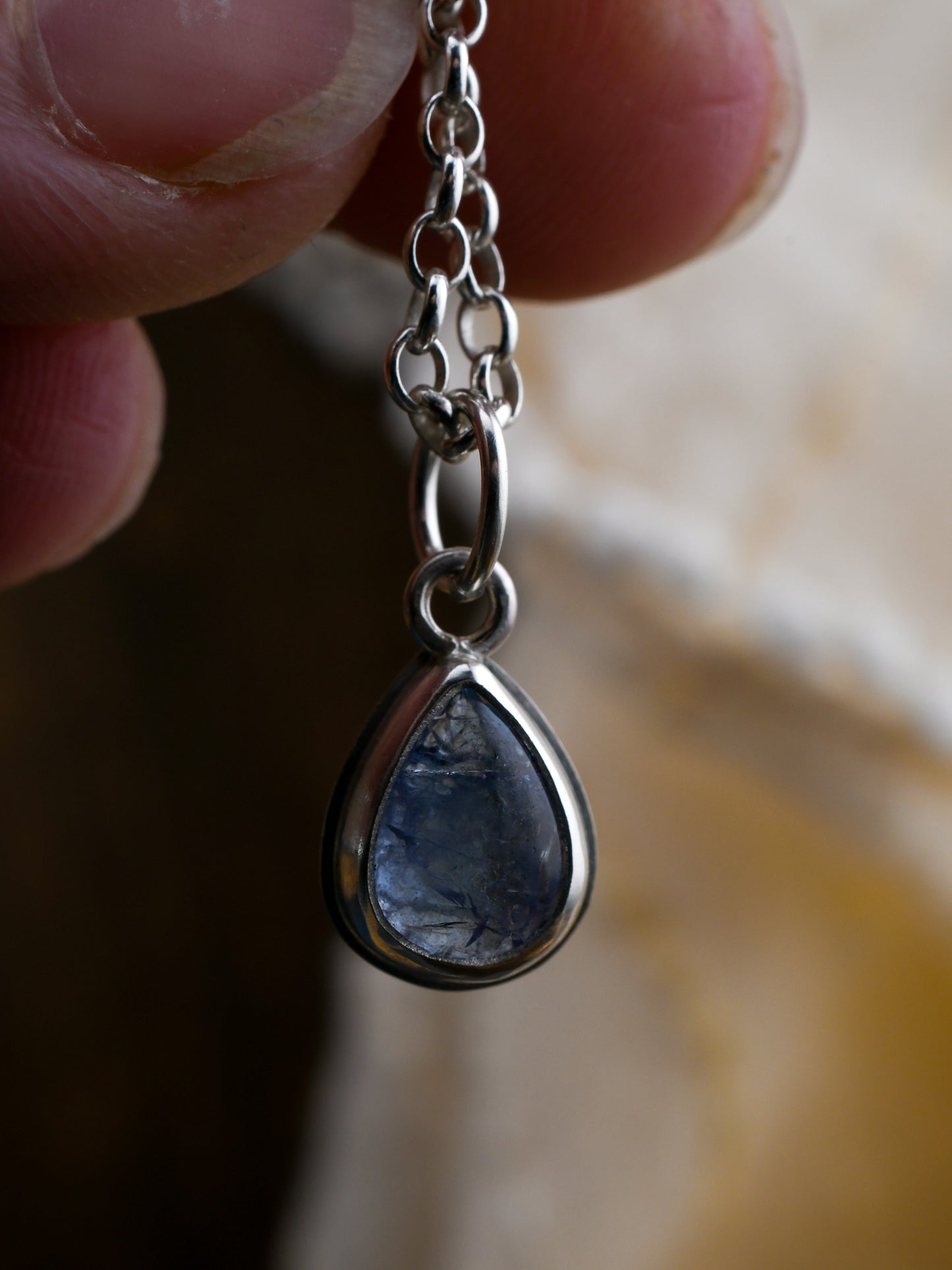 Teardrop-shaped blue stone pendant on a silver chain held by a hand.
