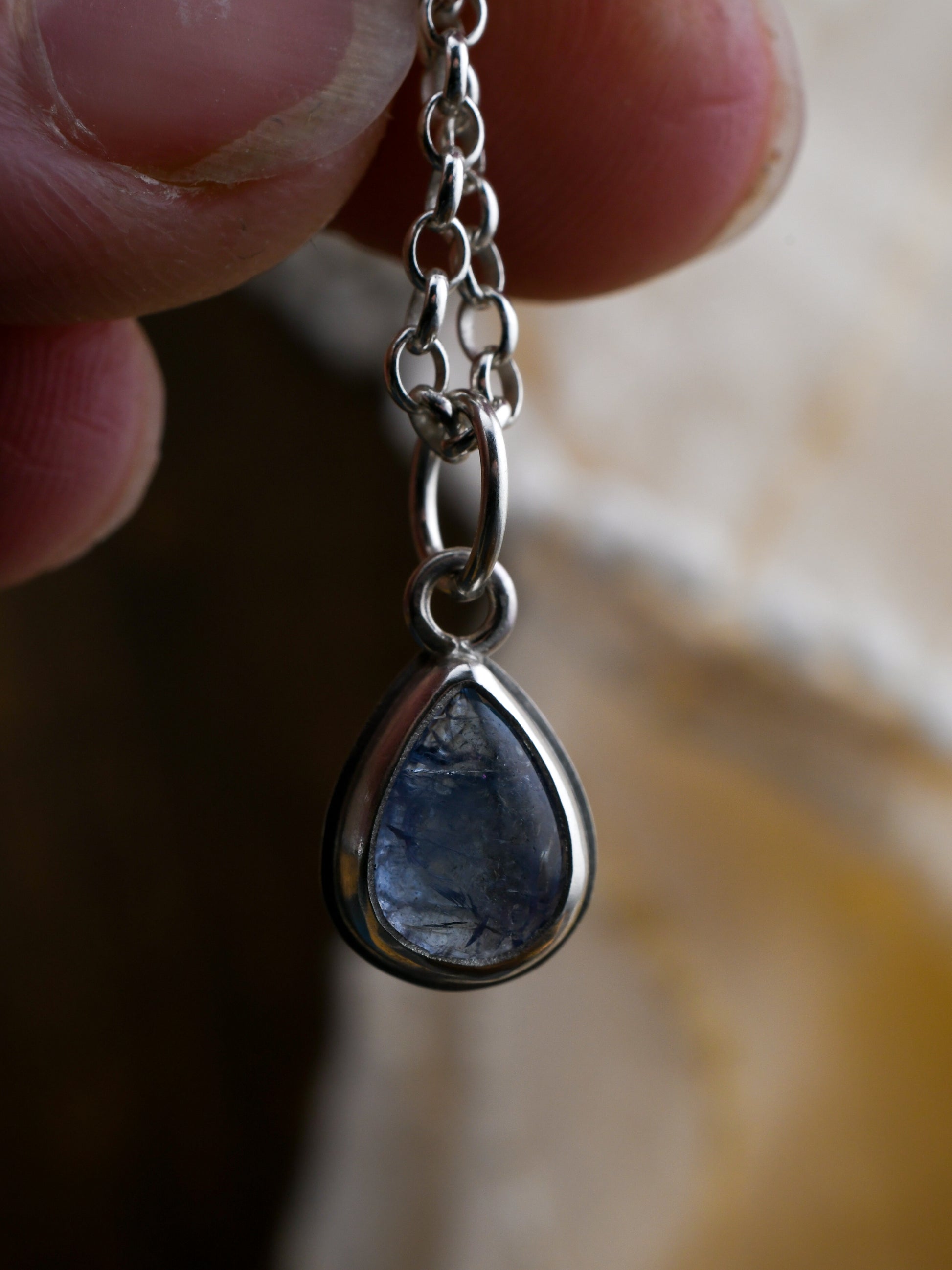 Teardrop-shaped blue stone pendant on a silver chain held by a hand.