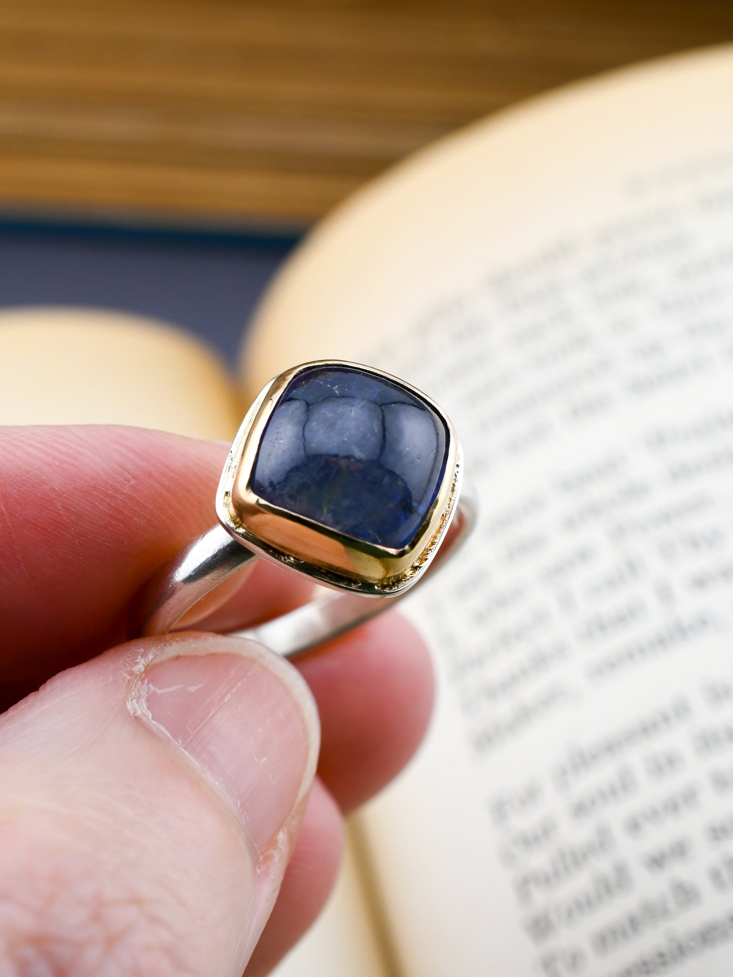 Gold and silver ring with a blue gemstone held by a hand against a blurred book background