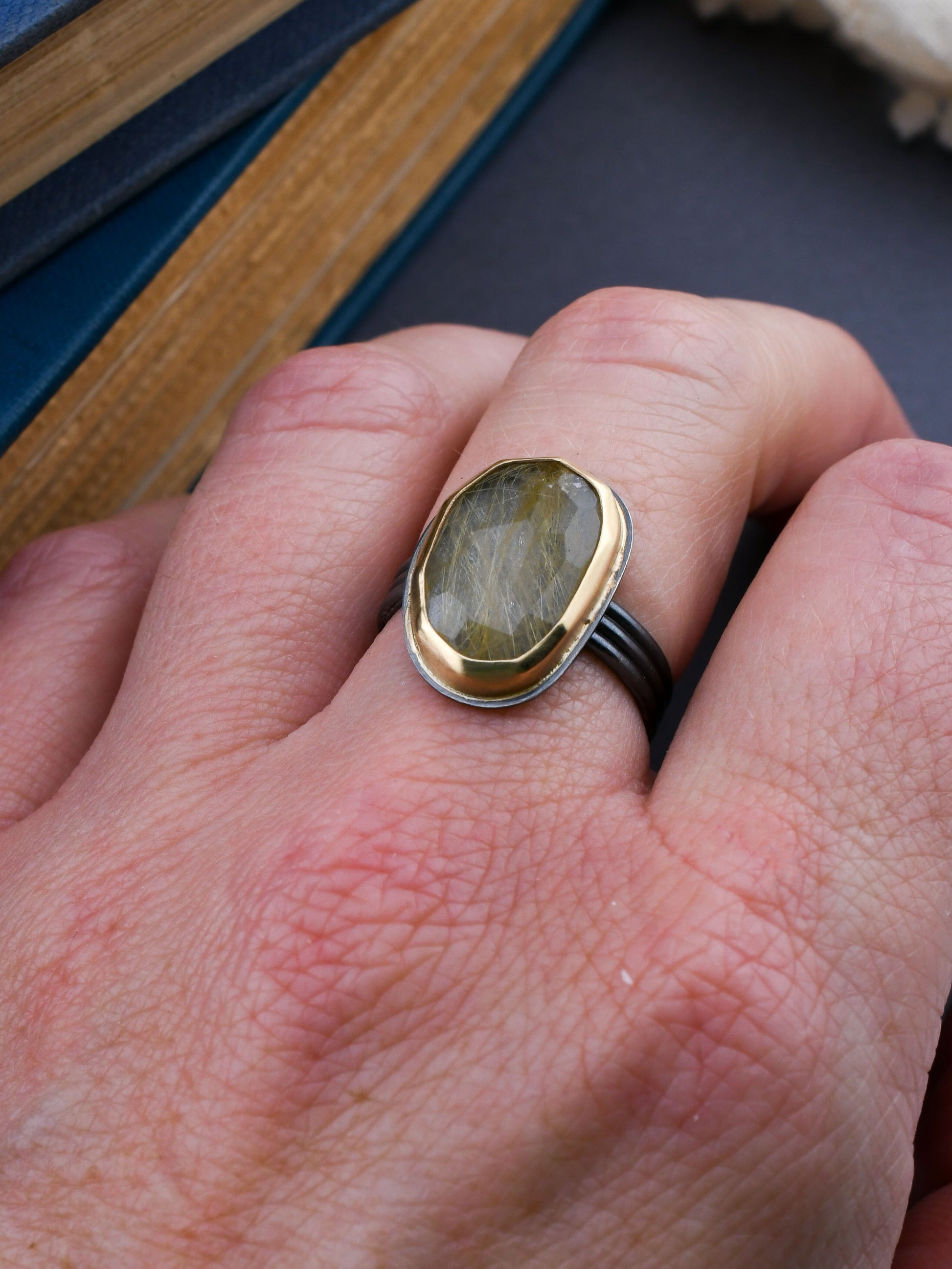 Hand wearing a ring with a golden rutilated quartz stone and gold bezel on a dark surface.