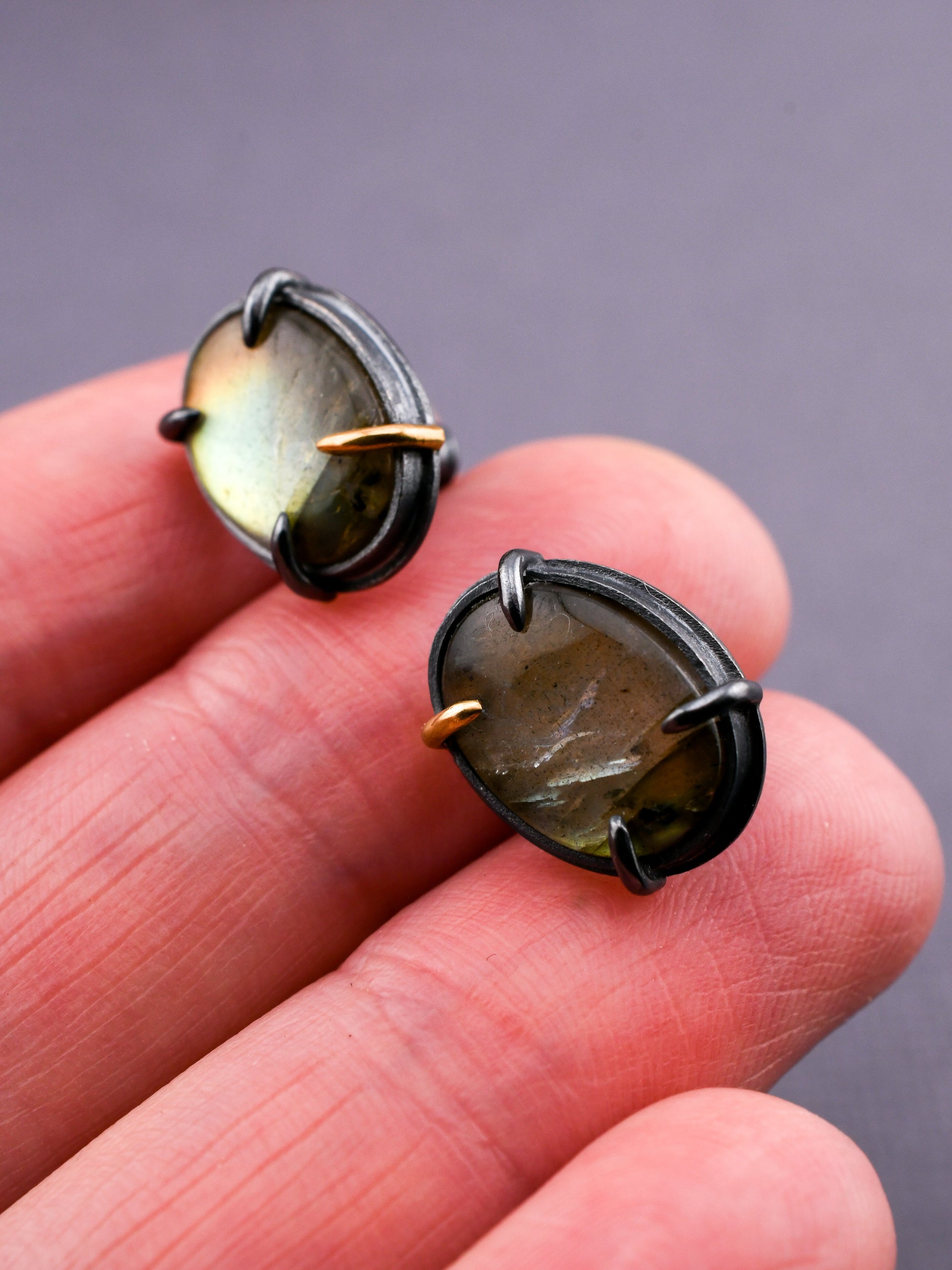 Close-up of two small round earrings with a metallic band on a hand against a gray background