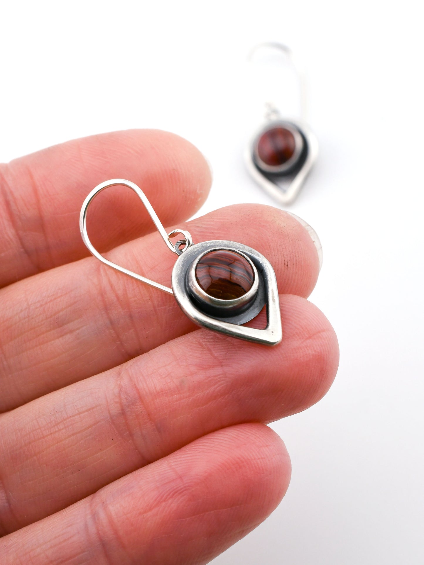 Silver earring with a banded red gemstone held between fingers on a white background
