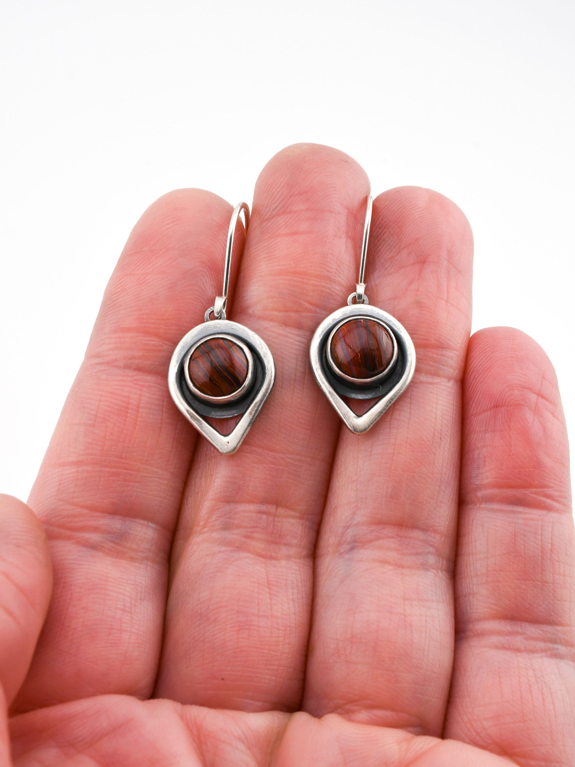 Silver earrings with banded red stones held between fingers on a white background