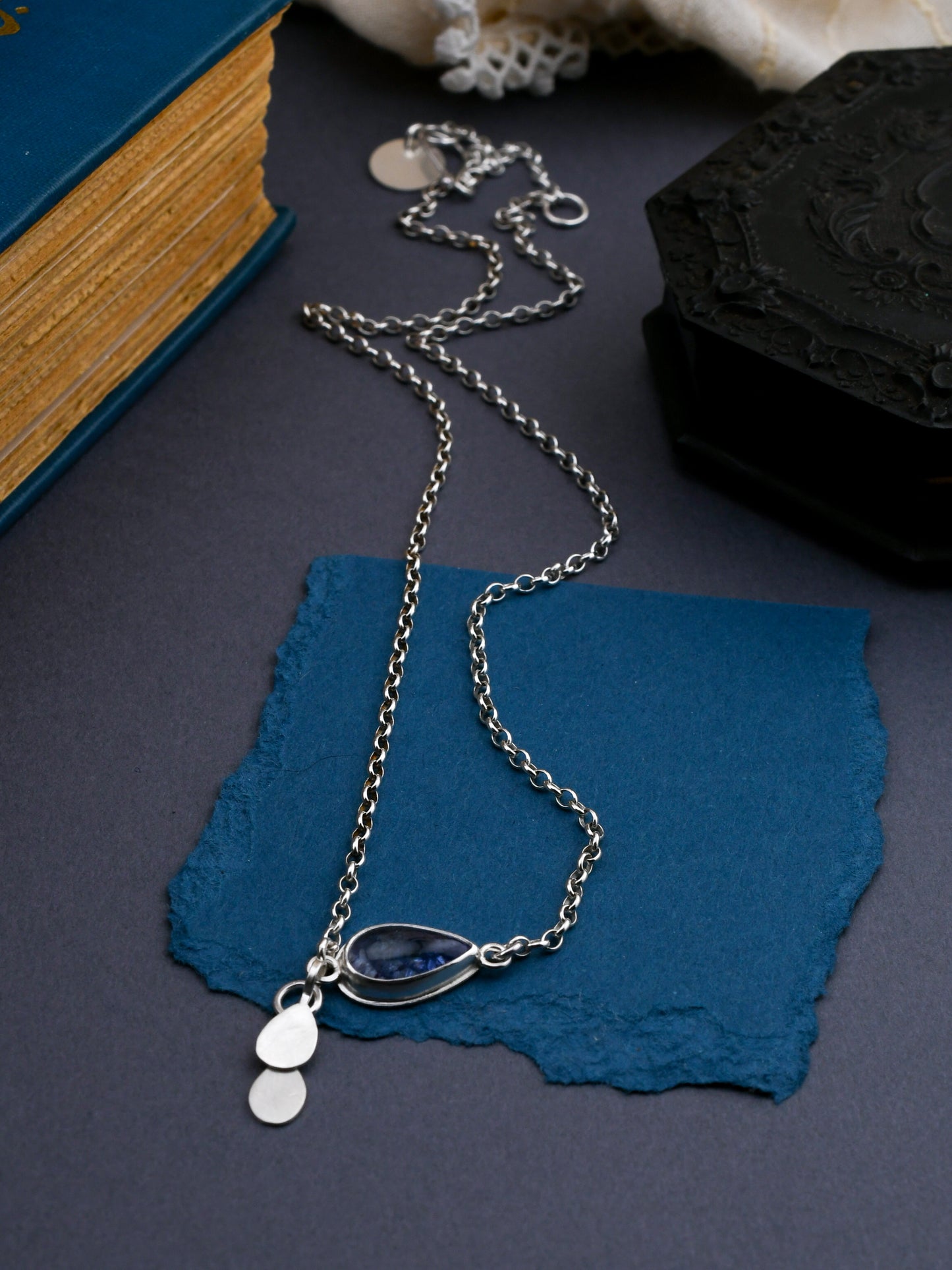 Silver necklace with pendant on blue paper, with a book and black box in the background