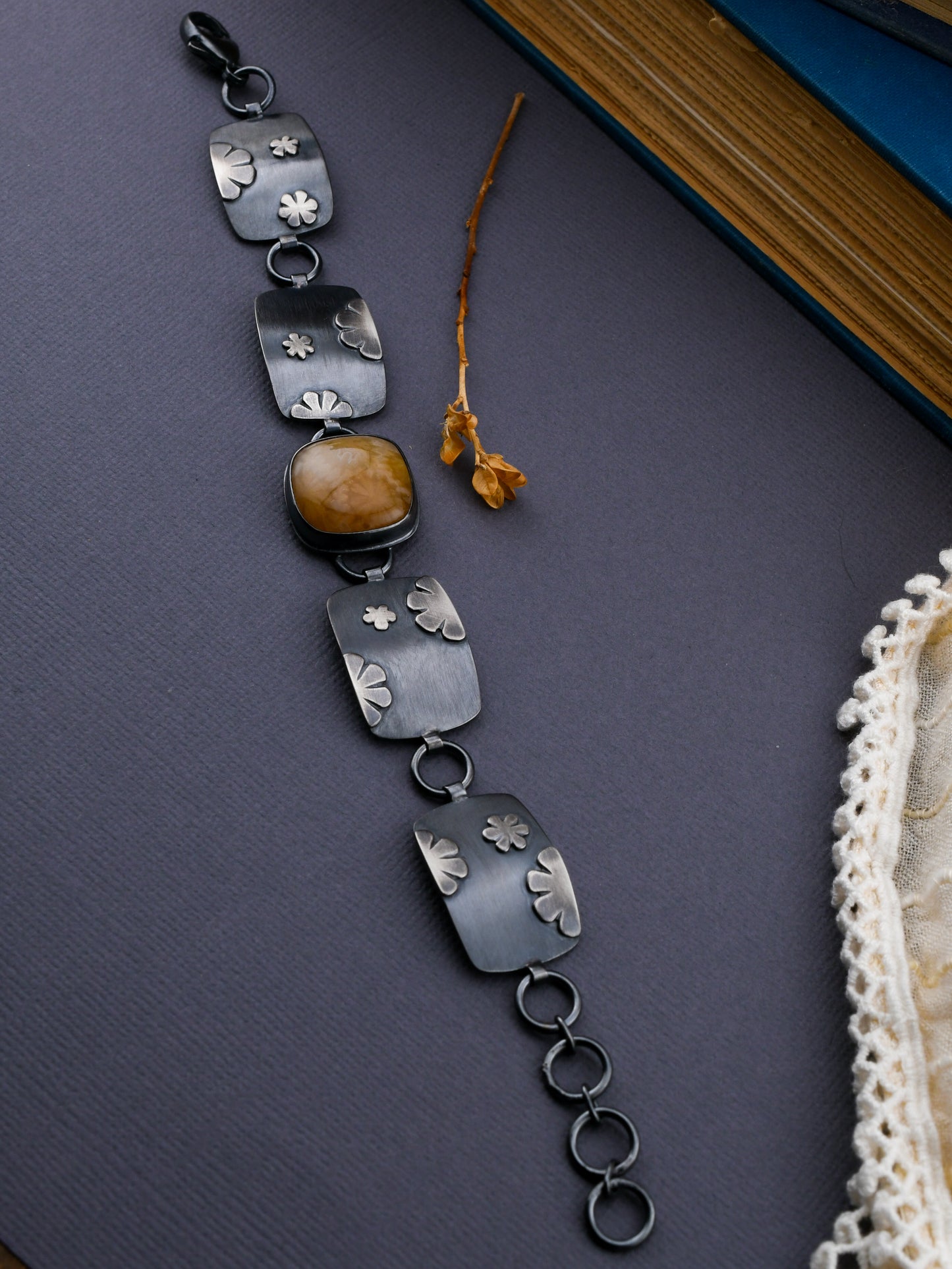 Black linked bracelet with gold-colored center stone on a gray background with dried flowers