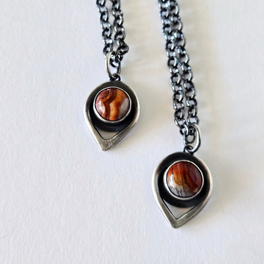 Two teardrop silver pendants with round crazy lace agate in red and orange patterns on a white background