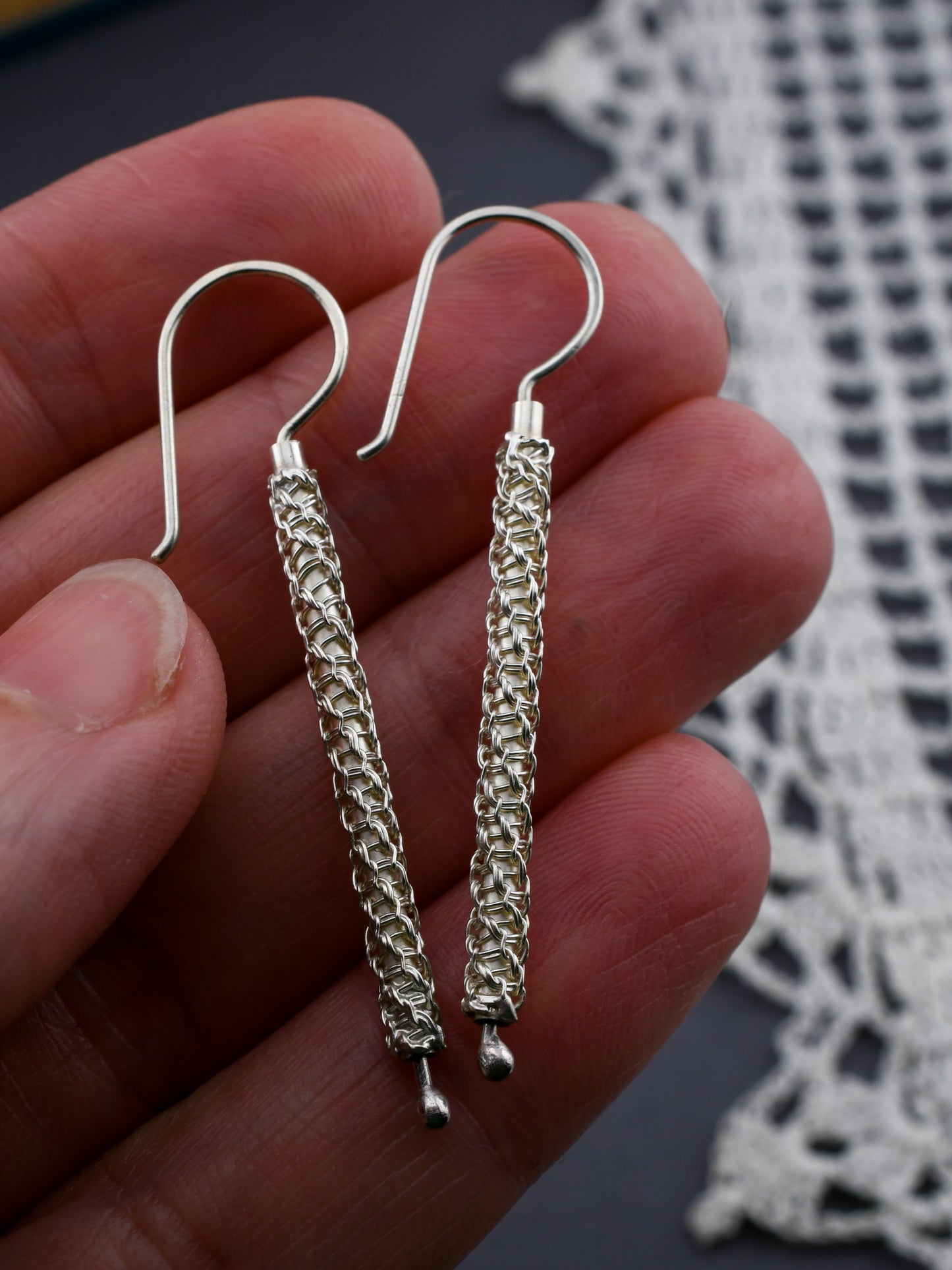Silver woven earrings held between fingers with a lace fabric background