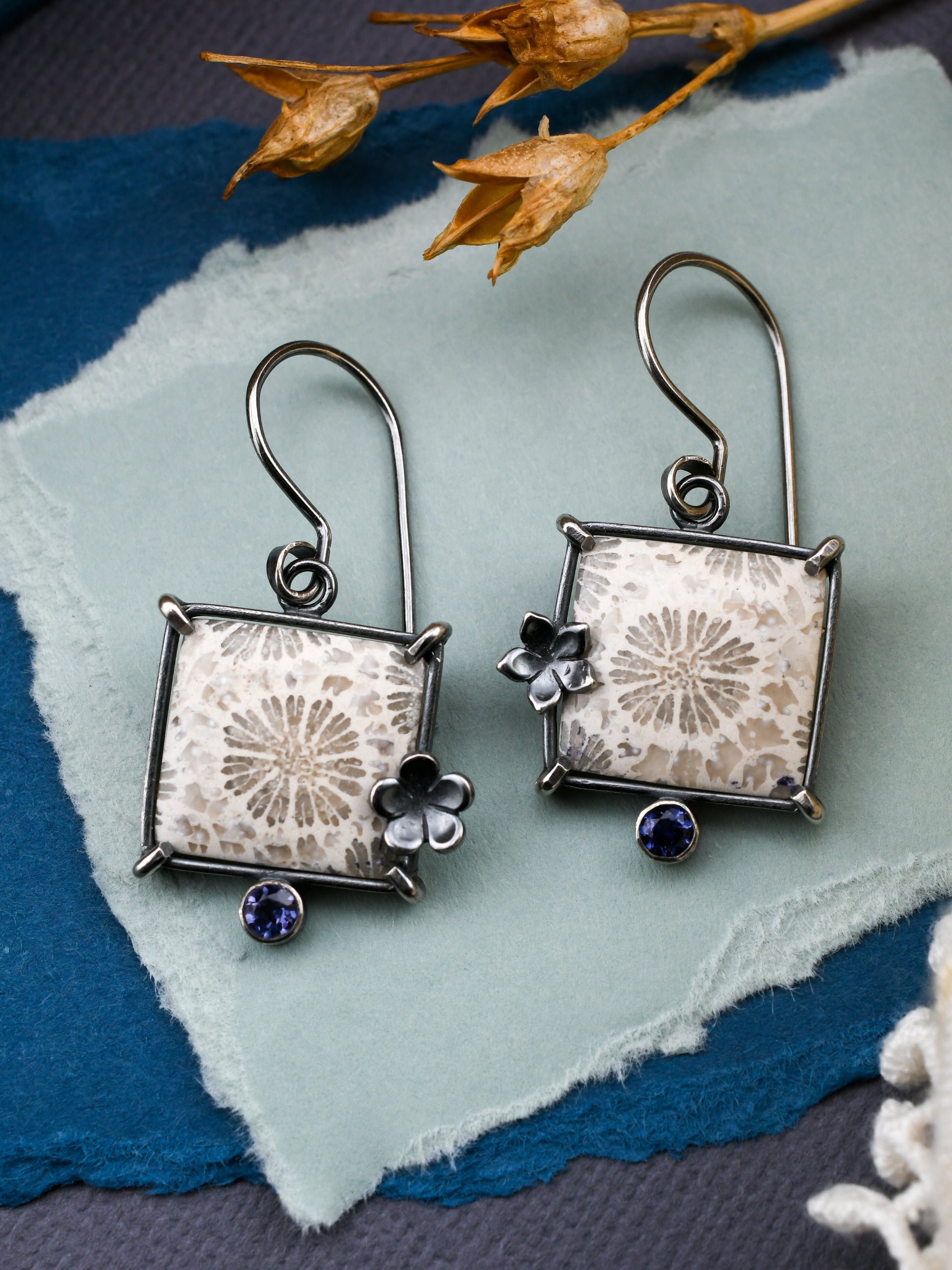 Silver earrings with square designs and violet stones on a textured blue surface.